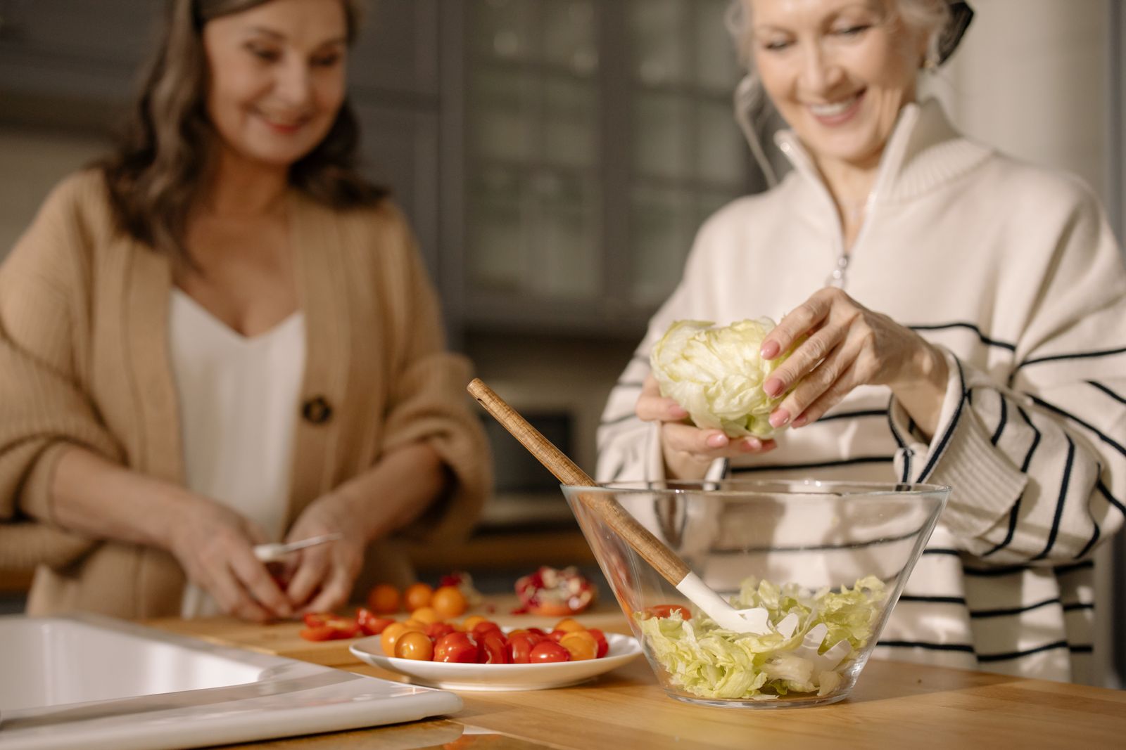 Two older women prepare a fresh salad together in a softly lit kitchen.