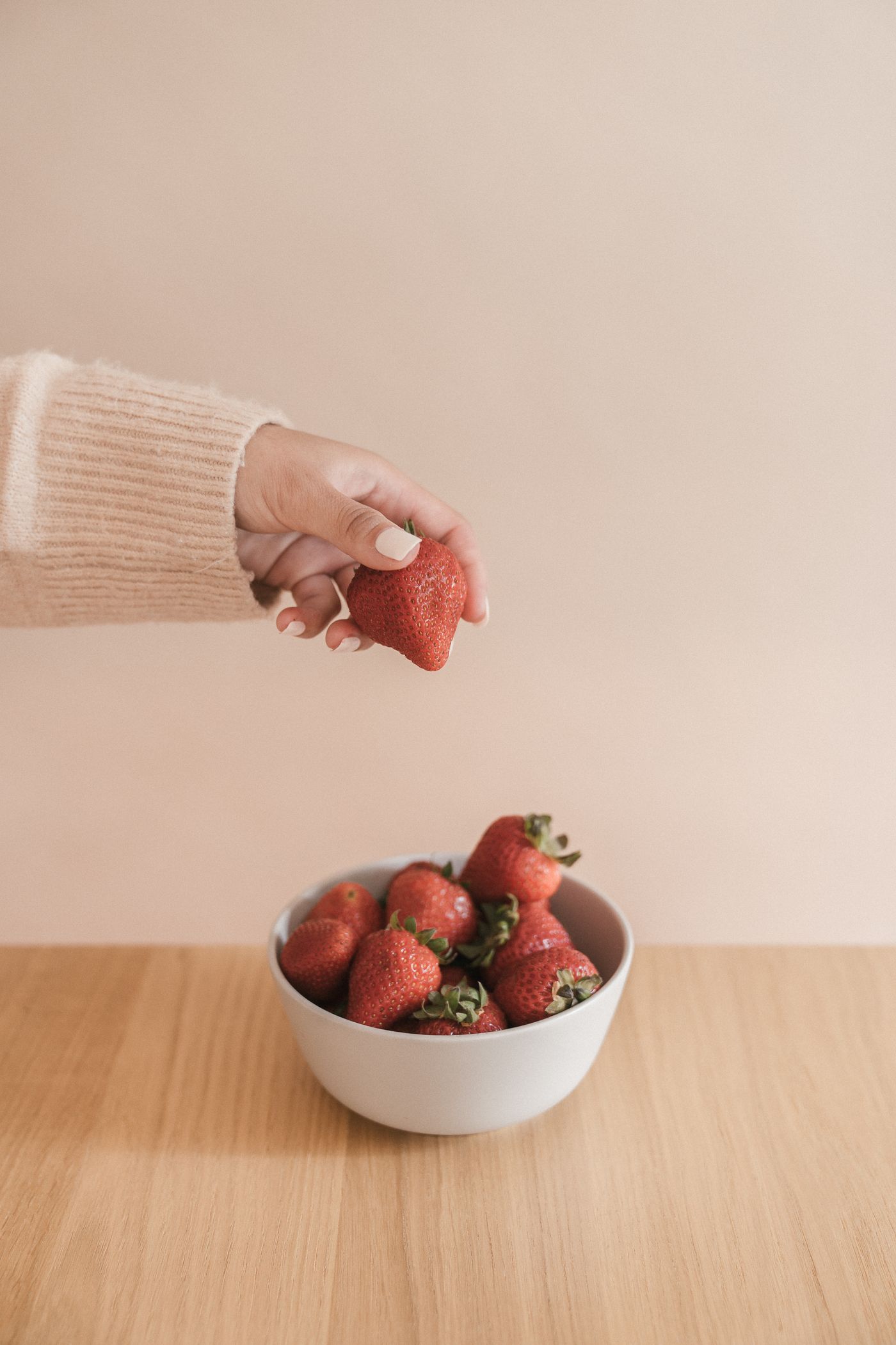 A hand reaches toward fresh strawberries in a bowl on a wooden table.